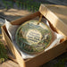 A decorative ornament in a box, featuring a scenic landscape with mountains, a waterfall, and a bridge.