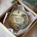 A decorative ornament in a box, featuring a scenic landscape of a lake surrounded by mountains and trees.
