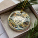 A decorative ornament in a box, featuring a scenic landscape with mountains, a cabin, and a stream.