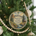 A round ornament hanging on a Christmas tree, featuring a scenic landscape with mountains, a river, and a cabin.