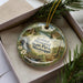 A decorative ornament in a box, featuring a scenic landscape with a waterfall, cabin, and pine trees.