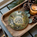 A decorative ornament featuring a scenic landscape with a waterfall, forest, and deer, placed on a wooden tray alongside pine cones and a lit candle.