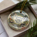 A decorative ornament in a box, featuring a scenic landscape with mountains, a river, and a bridge.