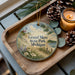 A decorative ornament with a lighthouse and a scenic landscape, placed on a wooden tray alongside pine cones and a lit candle.