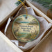 A circular ornament with a beach scene and a bird, placed in a box with a white tissue paper lining.