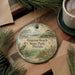 A circular ornament with a beach scene and a bird is displayed on a wooden surface, accompanied by a cup of coffee and some pine branches.