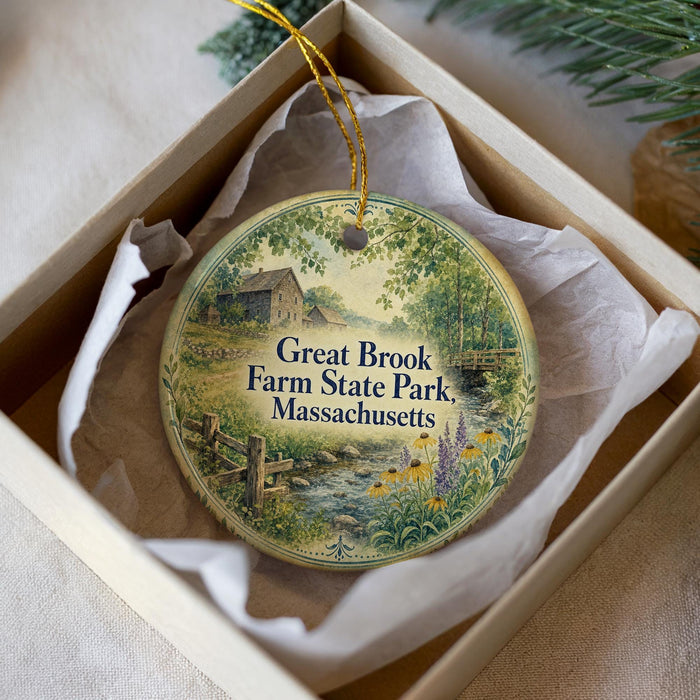 A round ornament with a painting of a farm and a stream, placed in a box with a white tissue paper lining.