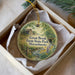A round ornament with a painting of a farm and a stream, placed in a box with a white tissue paper lining.