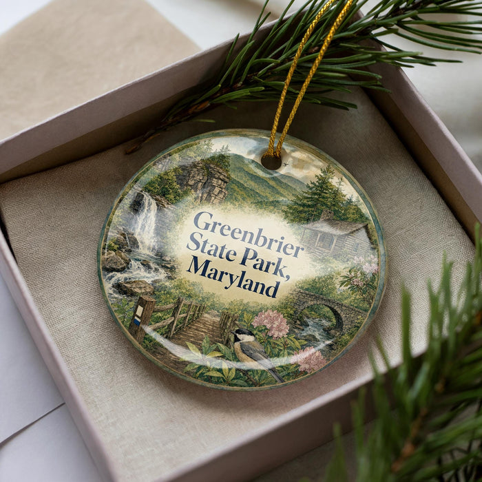 A decorative ornament in a box, featuring a scenic landscape with a waterfall, pine trees, and a bird.