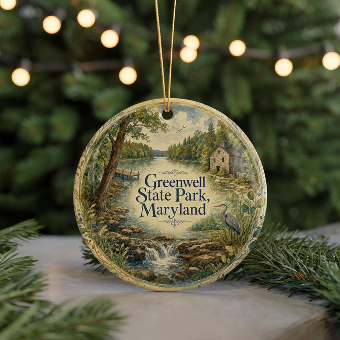 A circular ornament with a painting of a scenic landscape, featuring a river, trees, and a bird, hangs in front of a blurred Christmas tree with lights.