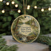 A circular ornament with a painting of a scenic landscape, featuring a river, trees, and a bird, hangs in front of a blurred Christmas tree with lights.