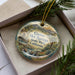 A decorative ornament in a box, featuring a scenic landscape with mountains, water, and greenery.