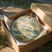 A circular ornament in a box, featuring a beach scene with a bird, a turtle, and a pier.