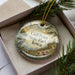 A decorative ornament in a box, featuring a scenic landscape with a bird, trees, and a body of water.