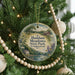 A round ornament hanging on a Christmas tree, featuring a scenic landscape with a house, trees, and a river.