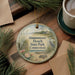 A round ornament with a beach scene and a bird on it, placed on a wooden surface next to a cup of coffee and some pine branches.
