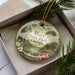A decorative ornament in a box, featuring a scenic landscape with a lake, trees, and a cabin, along with the text "Hart State Park, Georgia".
