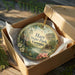 A decorative ornament in a box, featuring a scenic landscape with a cabin and flowers, with the text "Hart State Park, Georgia" printed on it.