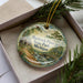 A decorative ornament with a scenic image of Hartwick Pines State Park, Michigan, displayed in a box with pine branches.
