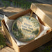 A round ornament in a brown box, featuring a scenic landscape with trees, flowers, and a body of water.