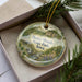 A decorative ornament in a box, featuring a scenic landscape with a river, trees, and mountains.