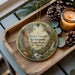 A wooden tray with a decorative ornament featuring a forest scene, pine cones, and a lit candle.