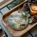A decorative ornament featuring a moose and a scenic landscape, with the text "Henry's Lake State Park, Idaho" displayed on it. The ornament is placed on a wooden tray, surrounded by pine cones and a lit candle.