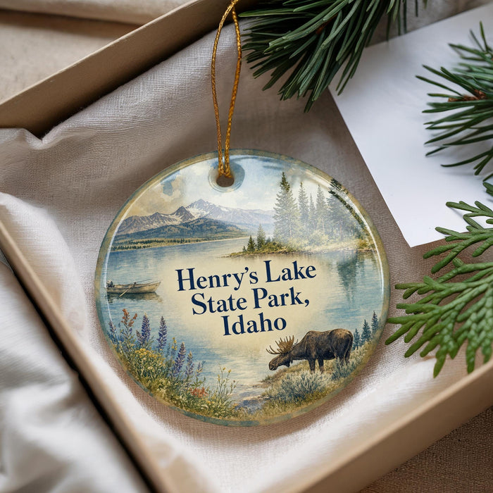 A decorative ornament with a moose and a scenic landscape, featuring mountains and a lake, is displayed in a box with pine branches.