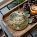 A decorative ornament featuring a scenic landscape with a deer, pine cones, and a candle on a wooden tray.
