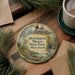A round ornament with a painting of a house and trees, surrounded by greenery, sits on a wooden surface.