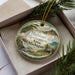A decorative ornament with a scenic landscape of a lake, mountains, and a cabin, hanging from a string.