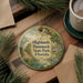 A round ornament with a scenic illustration of a park, featuring a bird, palm trees, and a pond, hangs on a wooden surface.