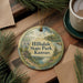 A decorative ornament with a scenic landscape of hills, water, and trees, featuring the text "Hillsdale State Park, Kansas" on it. The ornament is placed on a wooden surface, surrounded by pine branches and a cup of coffee.