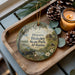 A decorative ornament with a painting of a historical site, specifically Historic Blakley State Park, Alabama, is displayed on a wooden tray alongside pine cones and a lit candle.