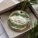 A decorative ornament in a box, featuring a scenic landscape with a deer, trees, and a waterfall.