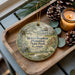 A decorative ornament featuring a house and greenery, placed on a wooden tray alongside pine cones and a lit candle.