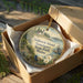 A round ornament in a box, featuring a scenic landscape with a lighthouse, trees, and a body of water.