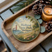 A wooden tray with a decorative ornament featuring a lighthouse and a scenic landscape, along with pine cones and a lit candle, creating a cozy and rustic atmosphere.