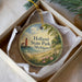 A round ornament with a lighthouse and beach scene, placed in a box with a white tissue paper lining.