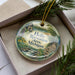 A decorative ornament in a box, featuring a scenic landscape with a lake, mountains, and a cabin.