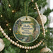 A round ornament hanging on a Christmas tree, featuring a scenic illustration of a lake, mountains, and a cabin.