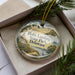 A decorative ornament in a box, featuring a scenic landscape with a lighthouse, trees, and a body of water.