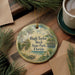 A round ornament with a tropical beach scene and palm trees, featuring the text "Hugh Taylor Birch State Park, Florida" on it, is displayed on a wooden surface alongside a cup of coffee and some pine branches.