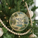 A round ornament hanging on a Christmas tree, featuring a tropical beach scene with palm trees, a house, and a bird.