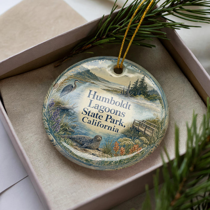 A decorative ornament with a scenic image of a lagoon, featuring a bird, a dock, and a body of water, is displayed in a box with pine branches.
