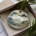 A decorative ornament with a scenic image of a lagoon, featuring a bird, a dock, and a body of water, is displayed in a box with pine branches.