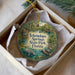 A circular ornament with a painting of a manatee and a bird, surrounded by a natural scene, is displayed in a box.