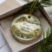 A decorative ornament in a box, featuring a scenic landscape with a bridge, trees, and a deer.