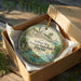 A circular ornament in a wooden box, featuring an illustration of a tropical beach scene with palm trees, a bird, and a building.