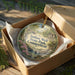 A circular ornament with a scenic landscape of Indian Well State Park, Connecticut, displayed in a wooden box with a gold string.